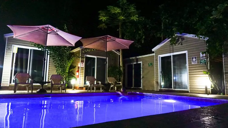 Piscina iluminada de noche con sombrillas y habitaciones al fondo