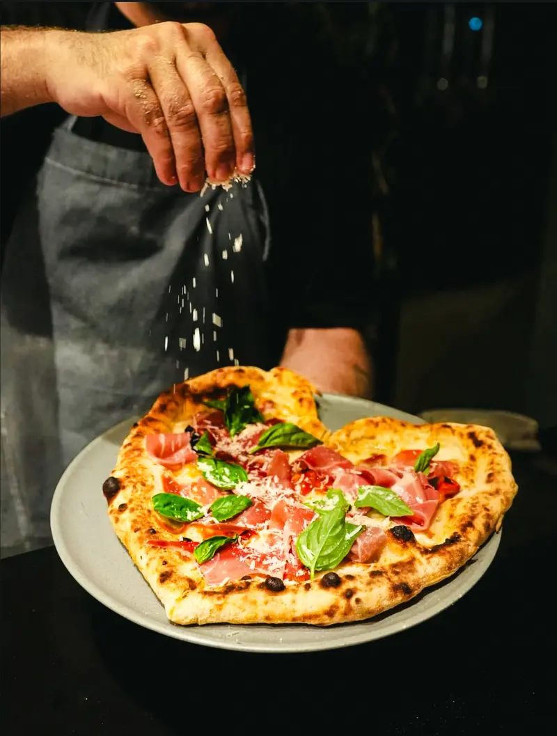 Pizza en forma de corazón con prosciutto crudo, albahaca y queso rallado mientras una mano espolvorea parmesano