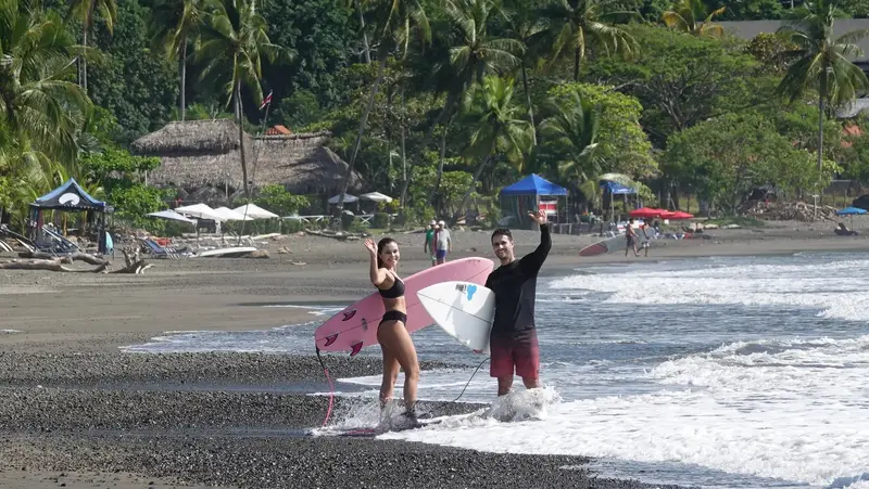 Una pareja sale del agua con sus tablas en una playa de arena oscura con palmeras