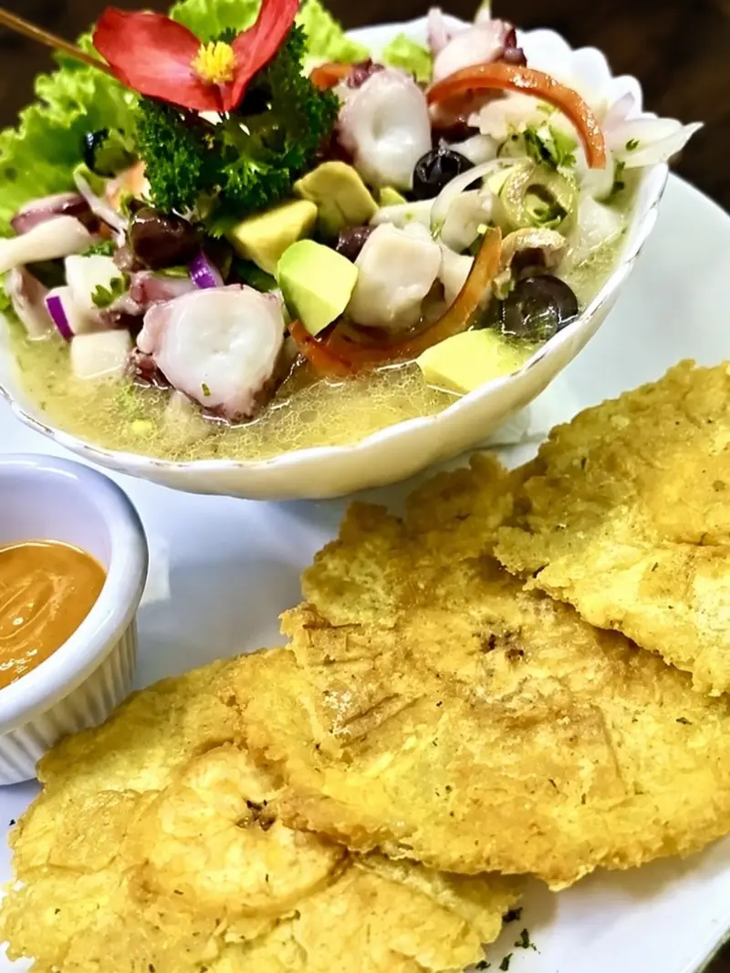 Ceviche mediterráneo con pulpo, aguacate y aceitunas en un tazón blanco, con patacones y una salsa al lado