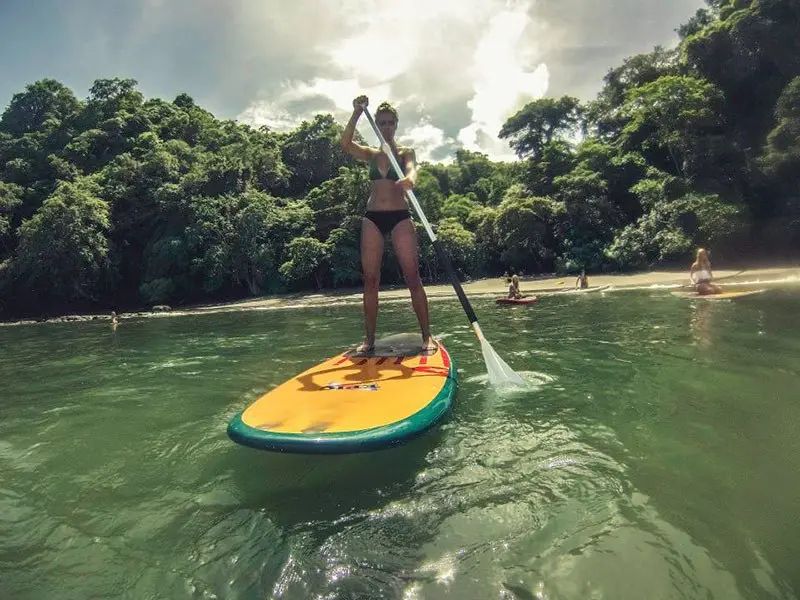 Persona remando de pie sobre una tabla de paddle amarilla en agua verde rodeada de selva